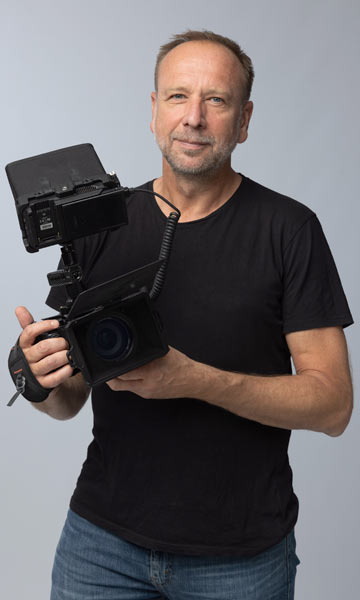 Videograf Hannover Udo Schlörmann steht mit der Filmkamera im Studio vor grauem Hintergrund und lächelt.